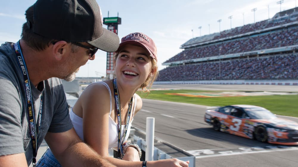 Father and teenage daughter enjoying a NASCAR race weekend from the stands as a stock car speeds by.