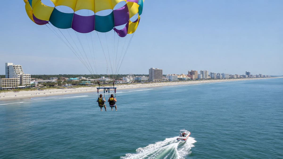 Two people parasailing above the ocean with the Myrtle Beach coast in view.
